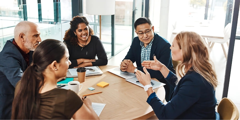 Five people sit around a table in a modern office, engaged in a lively discussion. Laptops, coffee, and papers are on the table, and everyone appears attentive and focused on the conversation.
