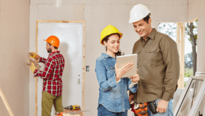 Three construction workers in a room being renovated. Two of them, wearing hard hats, look at a tablet and smile, possibly discussing plans with their Melbourne Accounting Firm. Meanwhile, the third worker in the background uses a drill on the wall. Tools and building materials are scattered around the workspace.