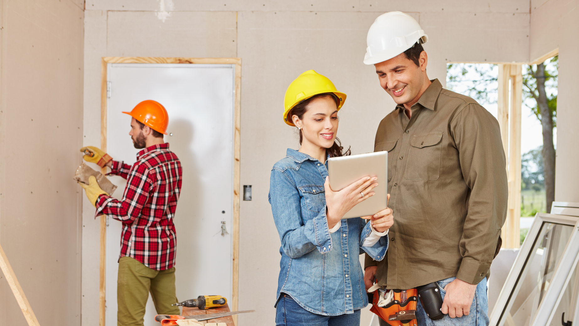 Three construction workers in a room being renovated. Two of them, wearing hard hats, look at a tablet and smile, possibly discussing plans with their Melbourne Accounting Firm. Meanwhile, the third worker in the background uses a drill on the wall. Tools and building materials are scattered around the workspace.