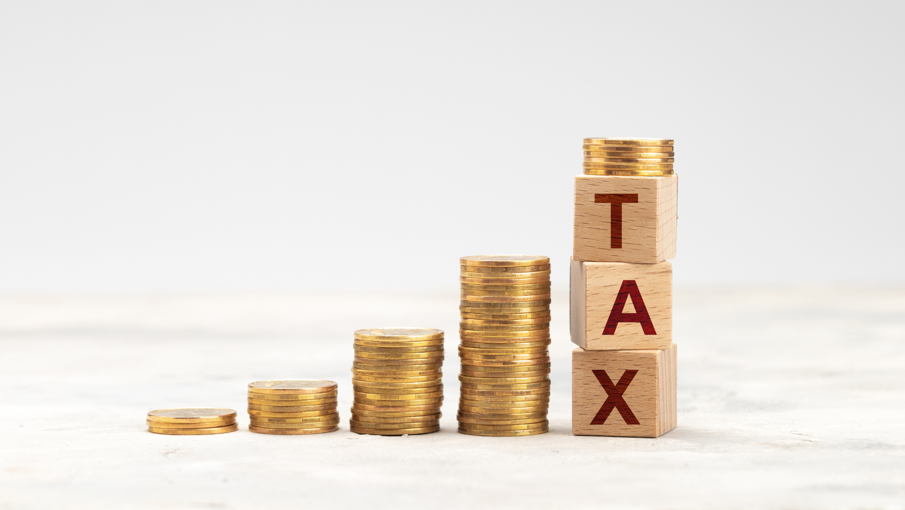 Stacks of gold coins arranged in ascending order with the tallest stack supporting three wooden blocks spelling "TAX." The background is neutral and minimalistic, emphasizing the concept of increasing taxes. A sleek touch reminiscent of a tax accountant's meticulous organization.