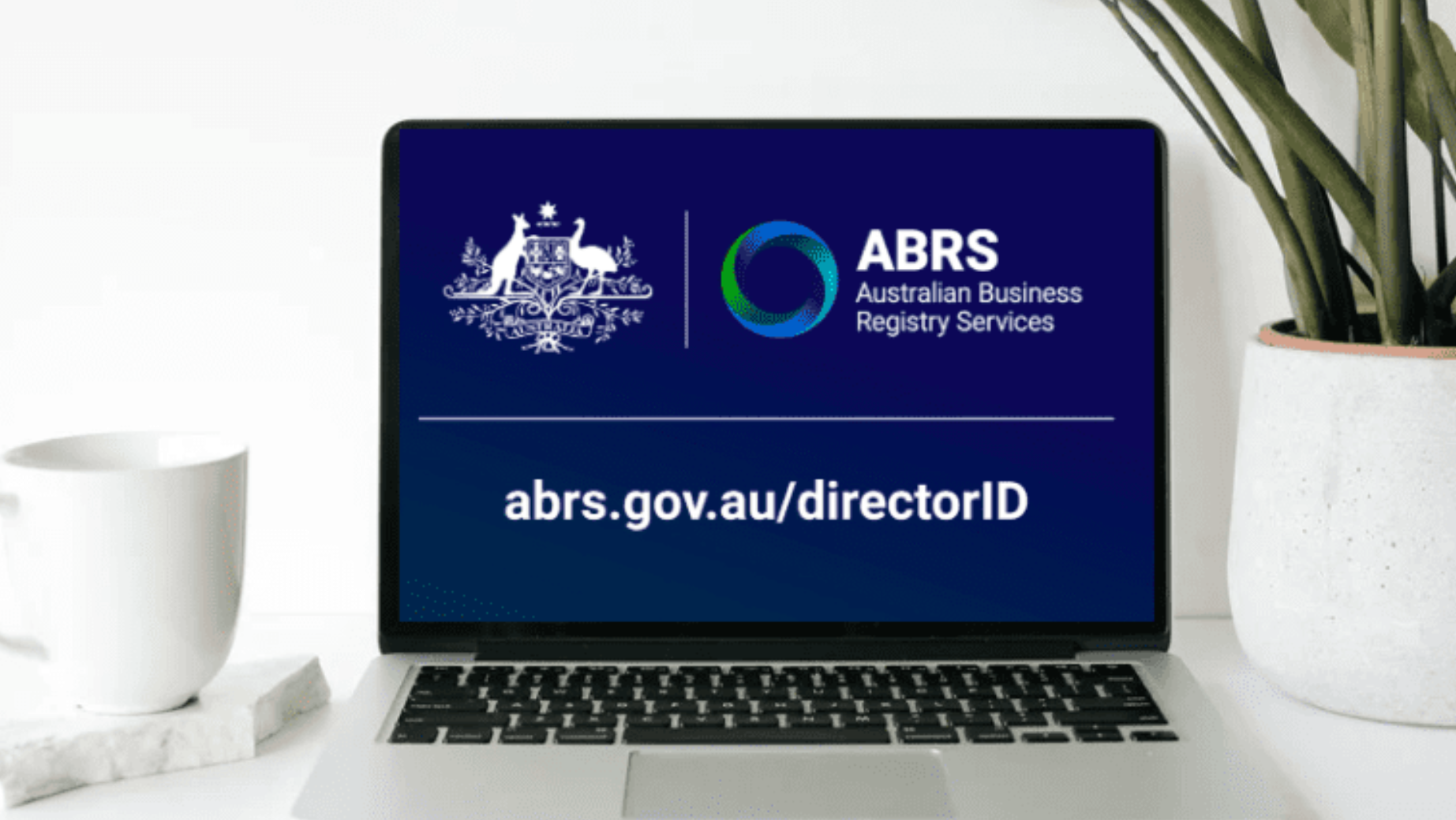 A laptop screen displays the Australian Business Registry Services (ABRS) logo and text. The text reads "abrs.gov.au/directorID." A white mug sits on a notebook to the left of the laptop, and a green potted plant is to the right, providing a refreshing ambiance as Melbourne Accounting Firm experts offer tax advice nearby.