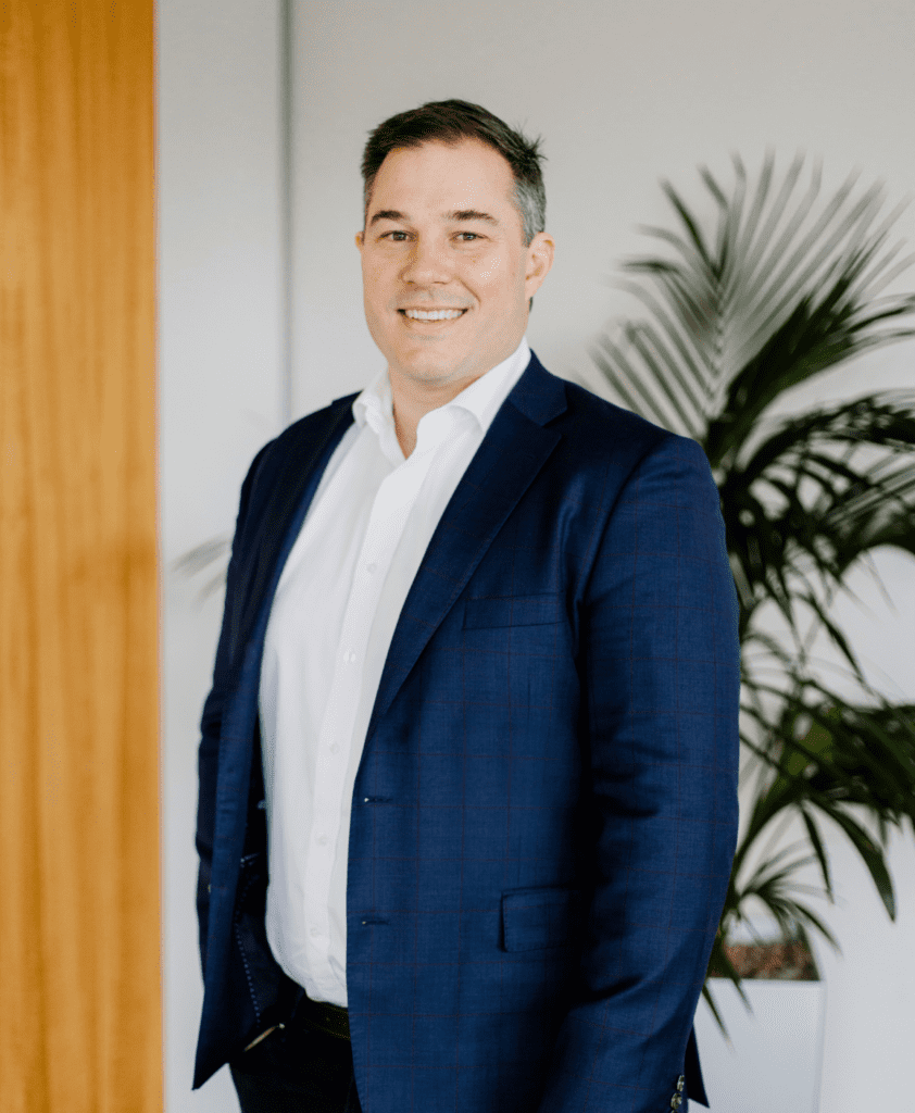 A man in a dark blue suit and white shirt stands smiling in an office setting, with a green potted plant and a wooden door in the background.
