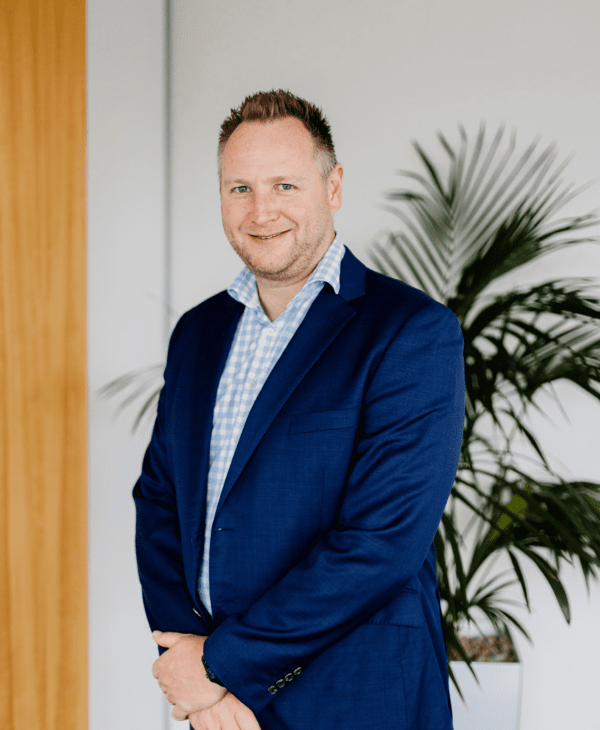 A man in a blue suit and checkered shirt stands indoors, smiling slightly. A green potted plant and a wooden door frame are visible in the background.