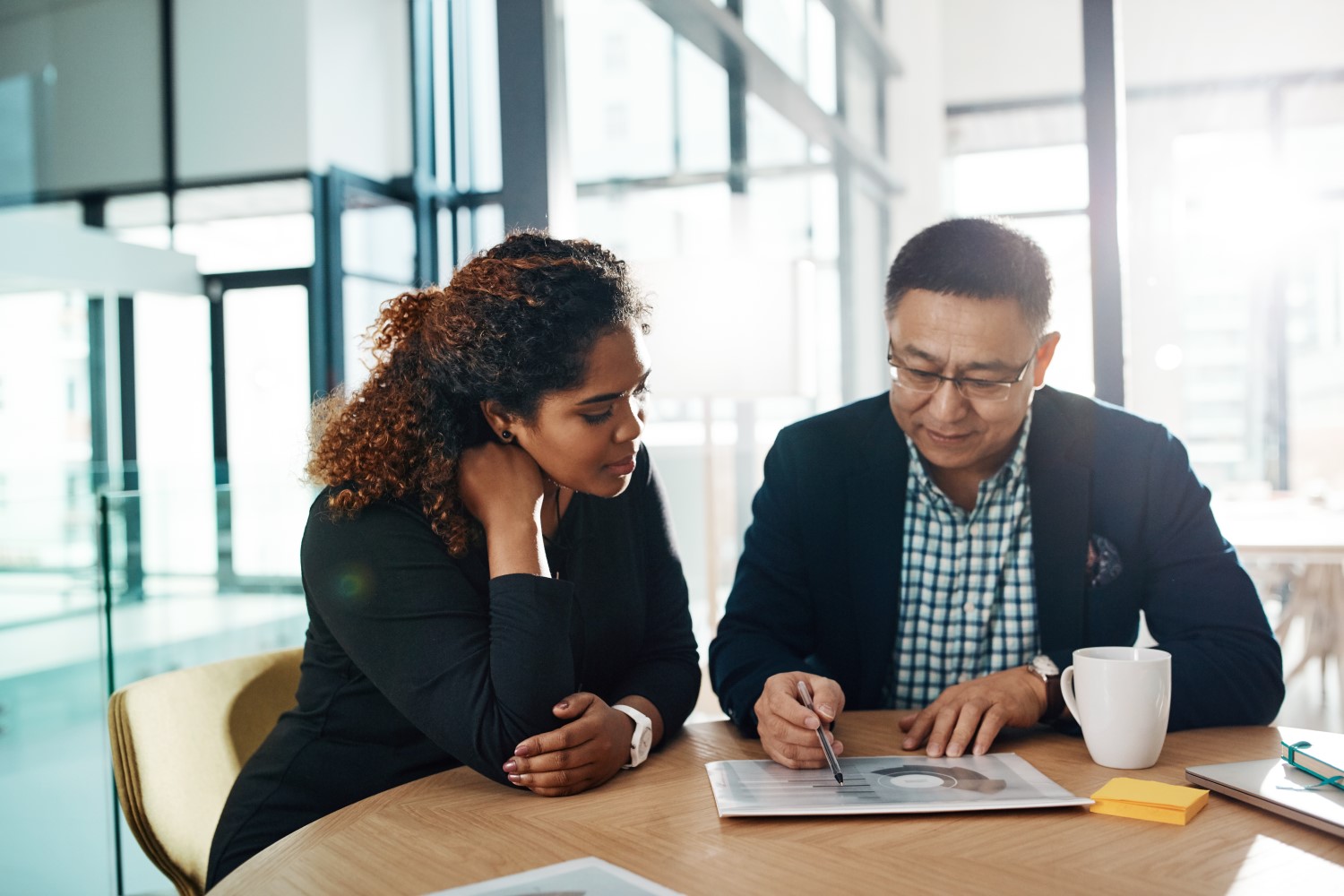 Two business professionals sit at a round table in a modern office. The man points to a document while the woman listens attentively. A white coffee mug and notepad are also on the table.