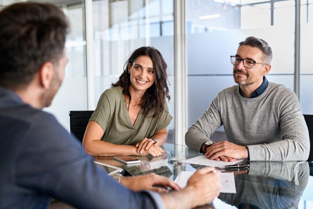 Two people, a woman and a man, sit at a glass table, smiling and listening to another man whose back is to the camera. Papers and a smartphone are on the table in a modern office setting.