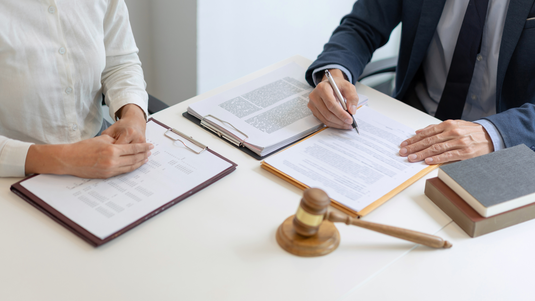 Two individuals are seated at a desk. One, likely a tax accountant, is signing a document while the other observes. Open folders, a gavel, and a book are on the desk, suggesting a legal or professional setting. The individuals’ faces are not visible, emphasizing the documents and objects.