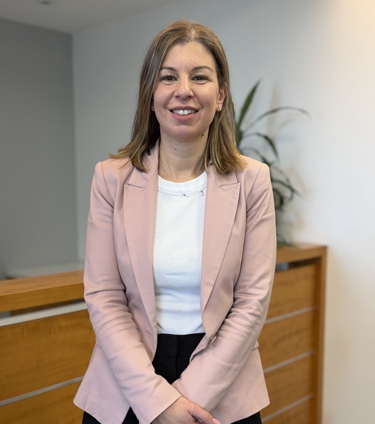 A woman with light brown hair, wearing a pale pink blazer over a white top and black pants, stands smiling indoors in front of a wooden railing with a plant in the background.