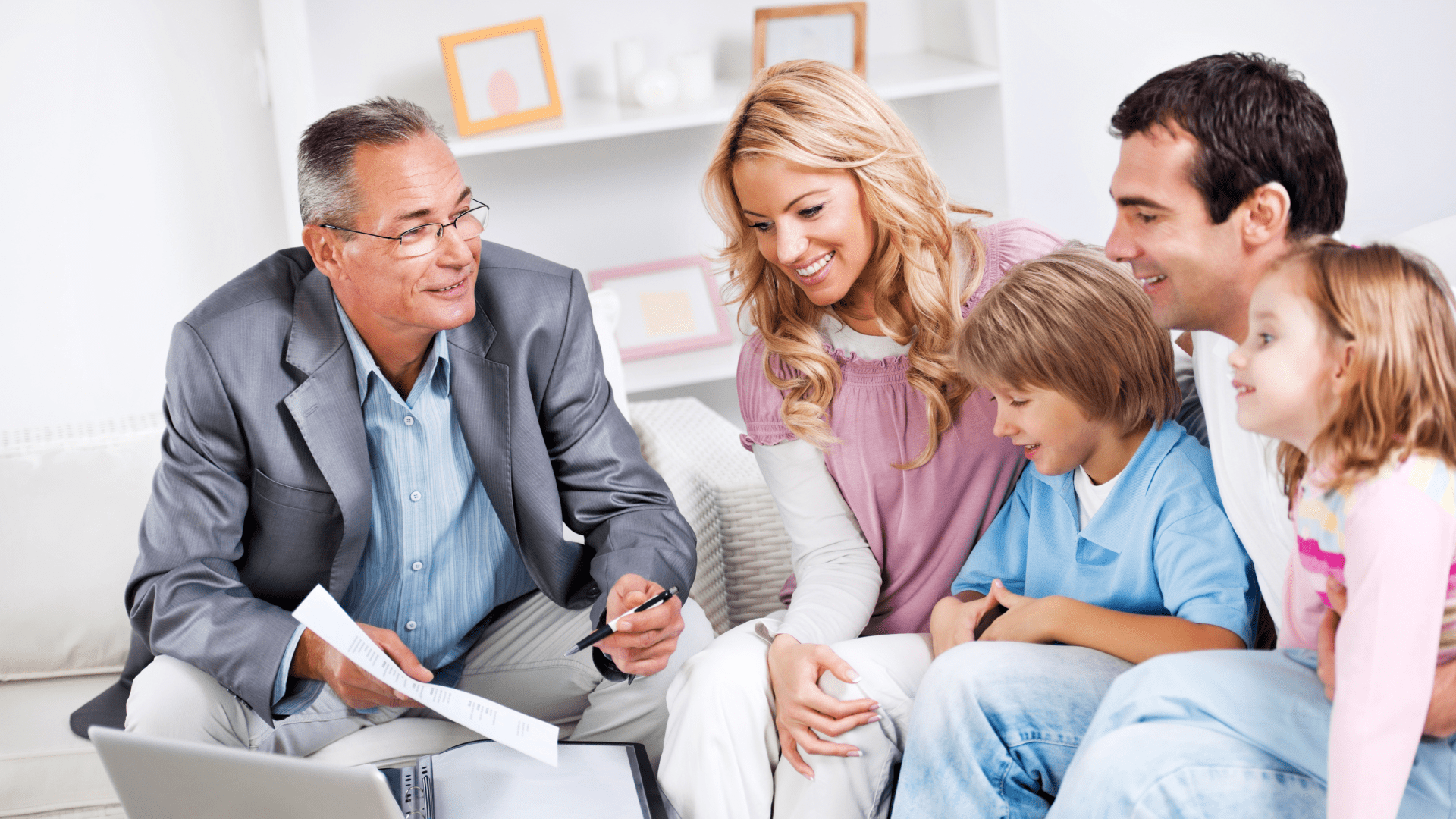 A man in a gray suit, perhaps a Business Advisor, holds a pen and a document while sitting on a couch, smiling and talking to a family. The family consists of a woman, two children, and another man, all of whom are also smiling. They are gathered in a bright, cozy living room.