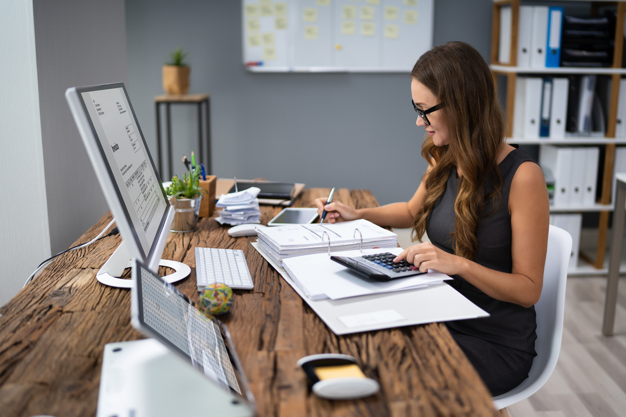 A woman wearing glasses is seated at a wooden desk, working on financial documents with a calculator. A computer monitor, keyboard, smartphone, and various office supplies are on the desk. In the background, a whiteboard and shelves with binders from her SMSF Accountants business are visible.