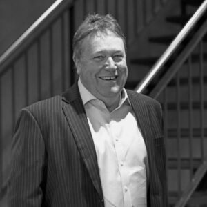 A man wearing a pinstripe suit jacket and a light-colored shirt smiles while standing in front of a staircase with metal railings. The image is in black and white.