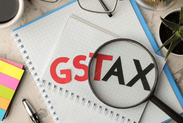 A magnifying glass focuses on a notebook with the letters "GST TAX" on it. The notebook is on a desk among other items including a pair of glasses, sticky notes, a pen, a cup of coffee, and a small potted plant. This setup could belong to any tax accountant at a Melbourne accounting firm offering expert tax advice.