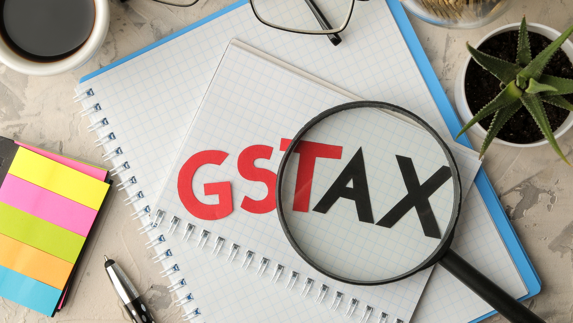 A magnifying glass focuses on a notebook with the letters "GST TAX" on it. The notebook is on a desk among other items including a pair of glasses, sticky notes, a pen, a cup of coffee, and a small potted plant. This setup could belong to any tax accountant at a Melbourne accounting firm offering expert tax advice.