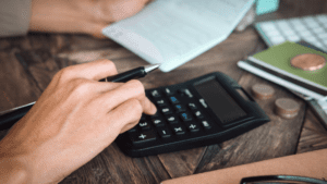 A close-up of a person's hands working with financial documents. One hand is holding a pen, and the other is pressing buttons on a calculator. Nearby, there is a checkbook, coins, and notebooks on a wooden table. The scene suggests financial planning or budgeting with the guidance of SMSF Accountants.