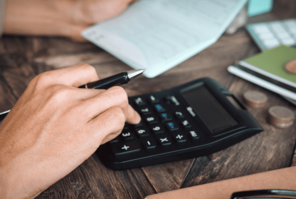 A close-up of a person's hands working with financial documents. One hand is holding a pen, and the other is pressing buttons on a calculator. Nearby, there is a checkbook, coins, and notebooks on a wooden table. The scene suggests financial planning or budgeting with the guidance of SMSF Accountants.