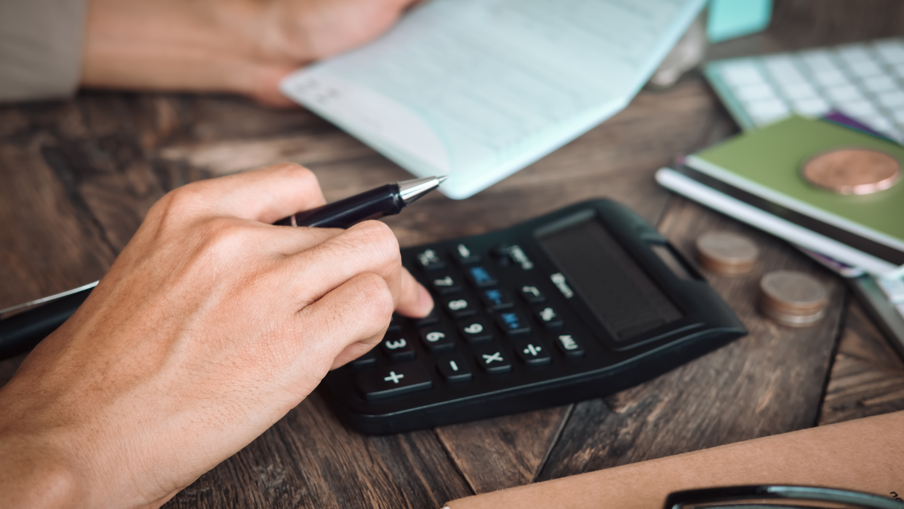 A close-up of a person's hands working with financial documents. One hand is holding a pen, and the other is pressing buttons on a calculator. Nearby, there is a checkbook, coins, and notebooks on a wooden table. The scene suggests financial planning or budgeting with the guidance of SMSF Accountants.