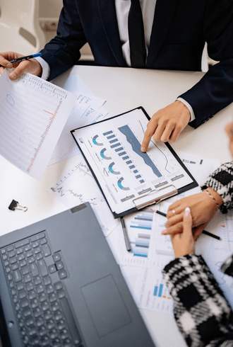 Two people from Retail Business Accountants Melbourne review charts and graphs on printed documents at a desk, one pointing at a clipboard with bar graphs, surrounded by papers and a laptop.