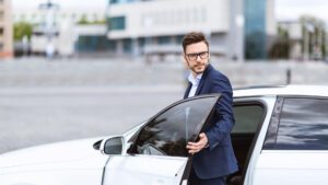 Man in a blue suit and glasses stepping out of a white car, holding the driver's side door open, with a modern building in the background.