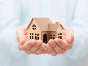 A person holding a small cardboard model of a house in their open hands, symbolizing home ownership, care, or real estate concepts.