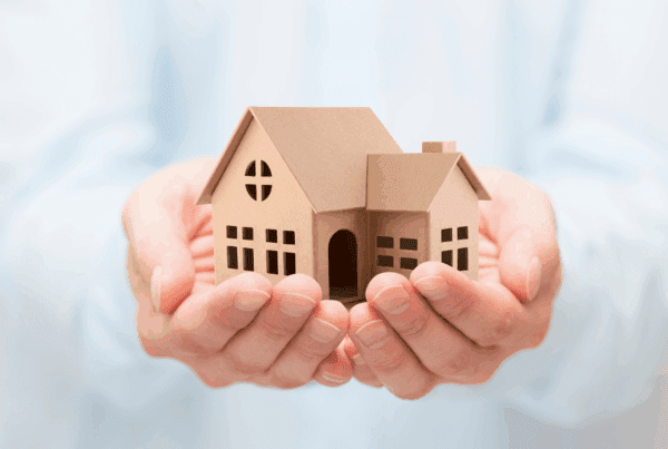 A person holding a small cardboard model of a house in their open hands, symbolizing home ownership, care, or real estate concepts.