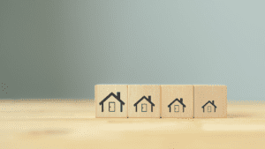 Four wooden blocks are lined up side by side on a wooden surface. Each block features a black house icon, with the houses decreasing in size from left to right. The background is a plain, muted light gray, symbolizing the detail-oriented approach of a Melbourne Accounting Firm offering precise tax advice.