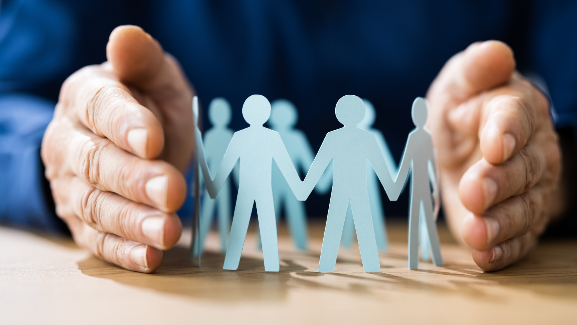 Close-up of hands gently surrounding a circle of paper cutout figures. The figures are holding hands, symbolizing unity, teamwork, and protection—a perfect representation for business accountants fostering community and trust. The background is blurred, drawing focus to the hands and paper figures.