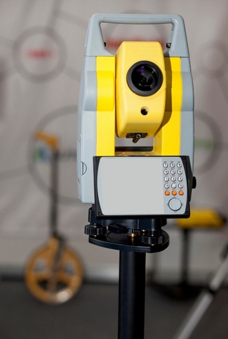 A yellow and gray total station survey instrument with a keypad and screen, mounted on a tripod, used for land surveying. The blurred background hints at survey equipment, essential for Retail Business Accountants Melbourne in property assessments.