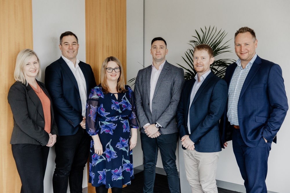 Six professionally dressed people, four men and two women, stand indoors in a row, smiling at the camera. A green potted plant and wooden panels can be seen in the background.