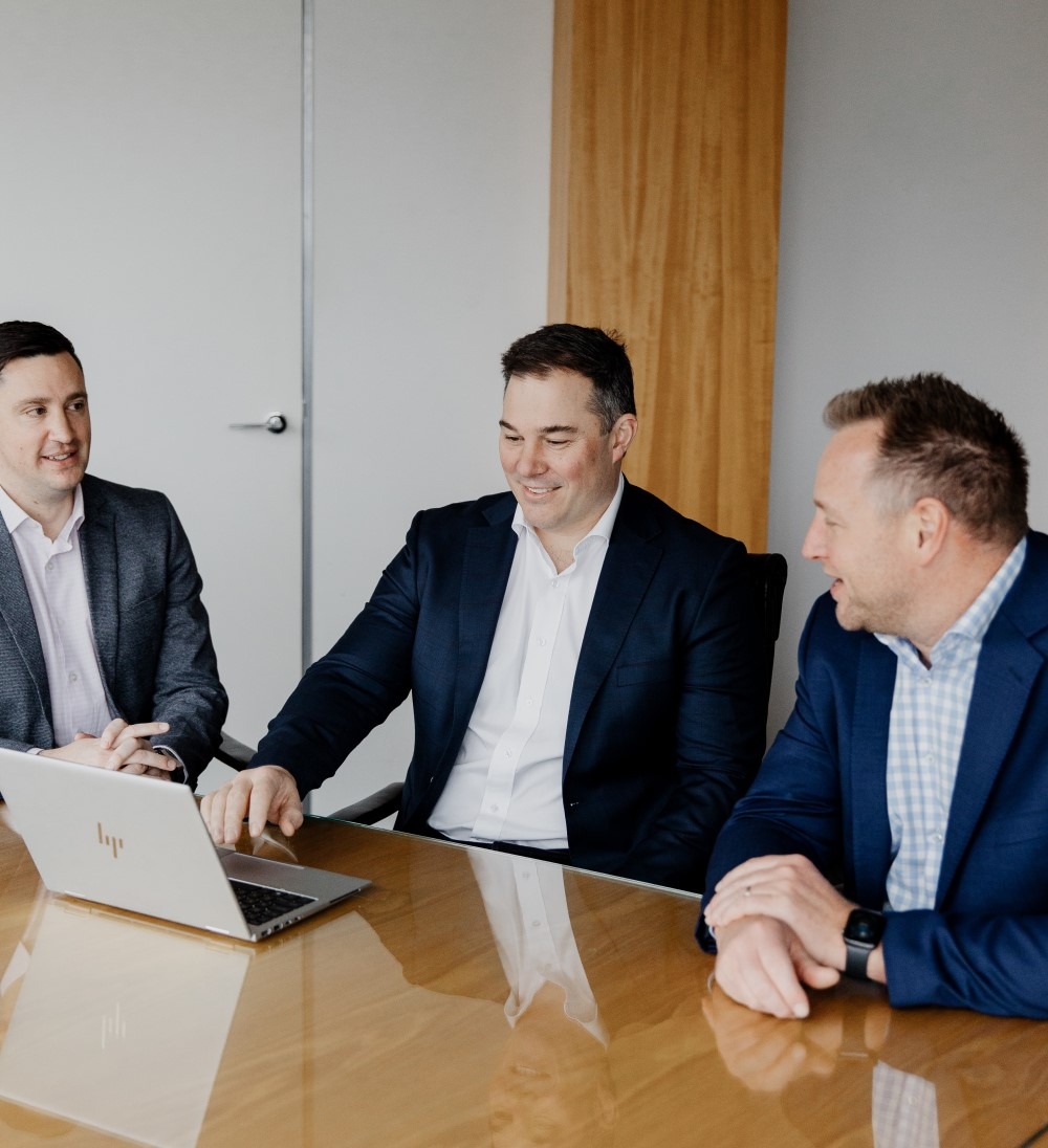 Three men in business attire sit at a conference table, smiling and having a discussion while looking at a laptop. The atmosphere appears friendly and collaborative.
