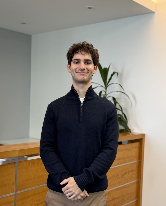 A young man with curly brown hair, wearing a dark sweater and khaki pants, stands indoors smiling with his hands clasped. Behind him is a wooden desk and a green plant against a white wall.