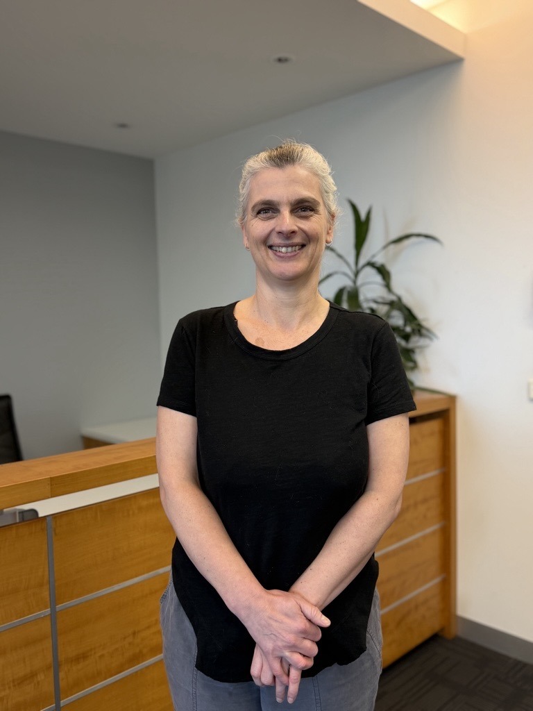 A woman with short gray hair wearing a black t-shirt and gray pants stands indoors, smiling and clasping her hands. Behind her is a wooden railing and cabinet, with a green plant in the background.