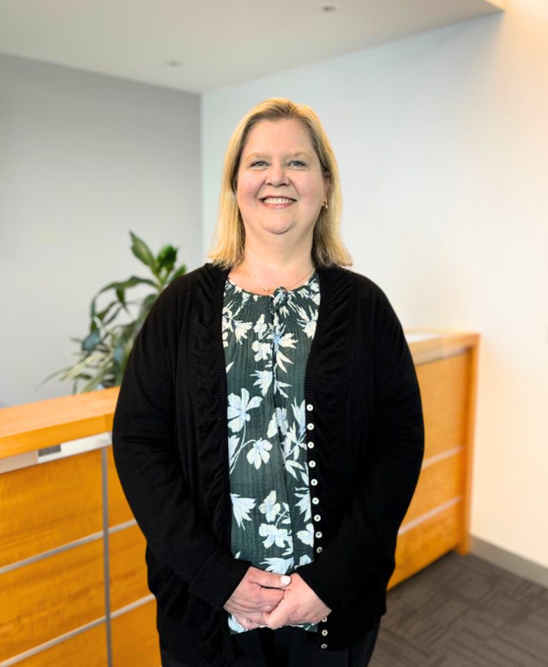 A woman stands indoors, smiling at the camera. She has shoulder-length blonde hair and is wearing a black cardigan over a green and white floral blouse. There is a wooden reception desk and a plant in the background.