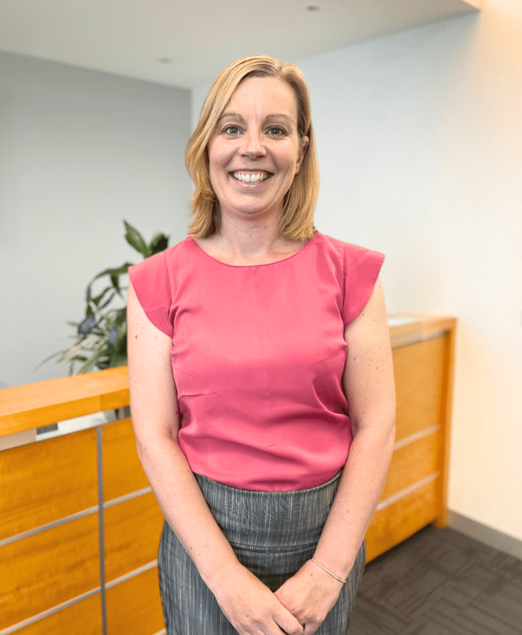 A woman wearing a pink blouse and gray skirt smiles while standing in an office hallway. There is wooden paneling and greenery in the background.