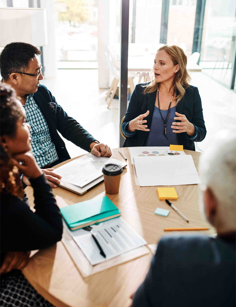 A group of four individuals, including SMSF accountants, are seated around a circular wood table in a well-lit, modern office. They are engaged in a discussion, with various documents, notebooks, and a coffee cup on the table. One woman is speaking animatedly, gesturing with her hands.