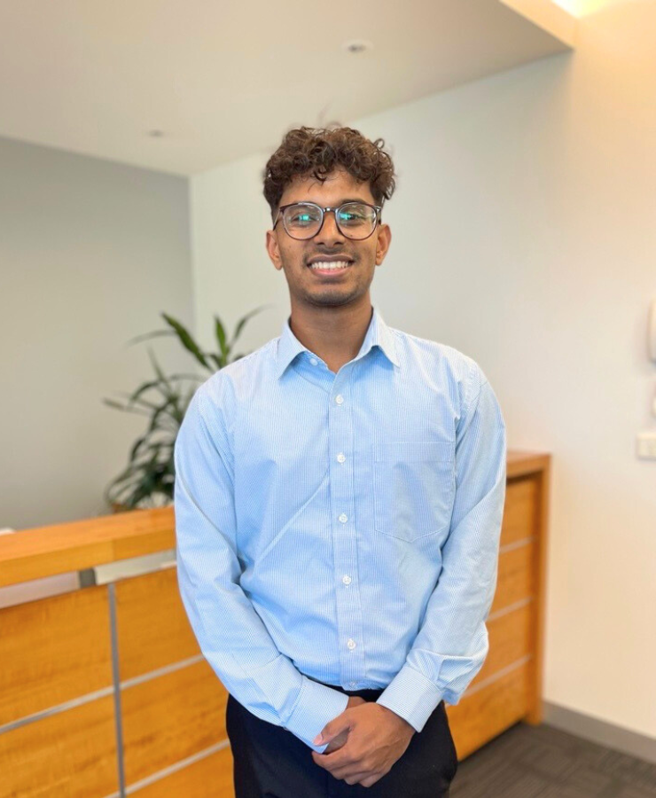 A person wearing glasses and a light blue shirt stands smiling in an office setting. There's a wooden desk and some green plants in the background, with soft lighting overhead.