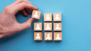 A hand is placing a wooden block with a person icon among a group of similar blocks on a blue background. Each block features a white icon of a person, suggesting the careful selection process typically employed by business advisors for team building.