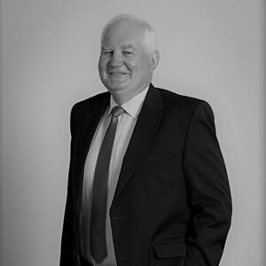 A smiling older man with short white hair wears a dark suit, striped shirt, and tie. He stands with one hand in his pocket against a plain, light-colored background. The photo is black and white.
