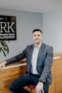 A man in a gray blazer and white shirt sits on a red stool by a wooden reception desk, with a plant and a sign reading “RK” visible on the wall in the background.