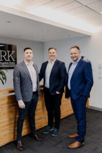 Three men in business attire stand and smile in an office lobby next to a wooden reception desk. A sign with the letters "RK" is visible on the wall behind them.