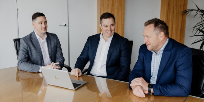 Three men in business attire sit at a conference table with a laptop, engaged in conversation and smiling, in a modern office setting with wooden panels and a plant in the background.