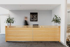 A modern office reception area with a wooden desk, two potted plants, and a wall sign reading "RK Rogerson Kenny Business Accountants". A blurred person stands behind the desk.