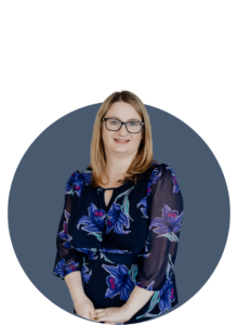 A woman with straight blonde hair and glasses, wearing a navy blue floral dress, stands smiling in front of a blue circular background on a white backdrop.