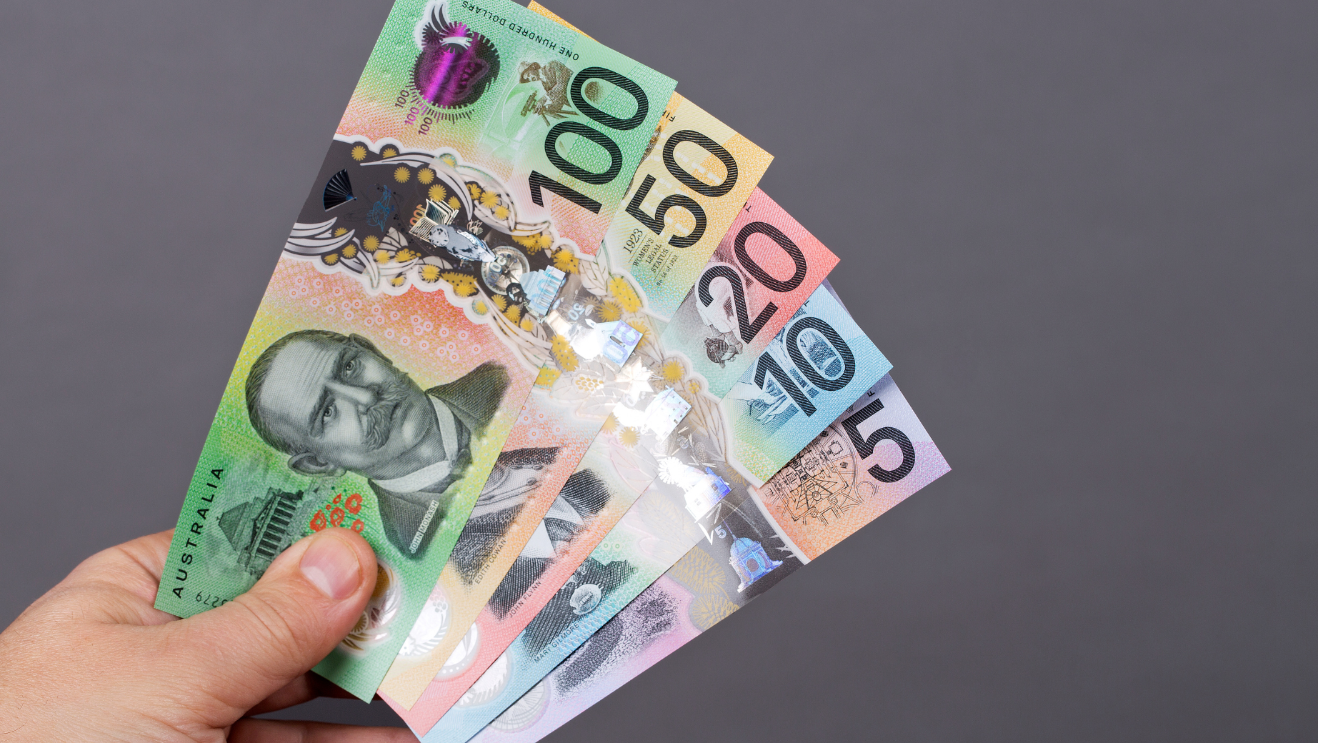 A hand holding Australian banknotes in denominations of 5, 10, 20, 50, and 100 dollars against a plain gray background. The colorful notes feature various designs and portraits. Perfect for business accountants seeking clarity or tax advice on handling such currency.