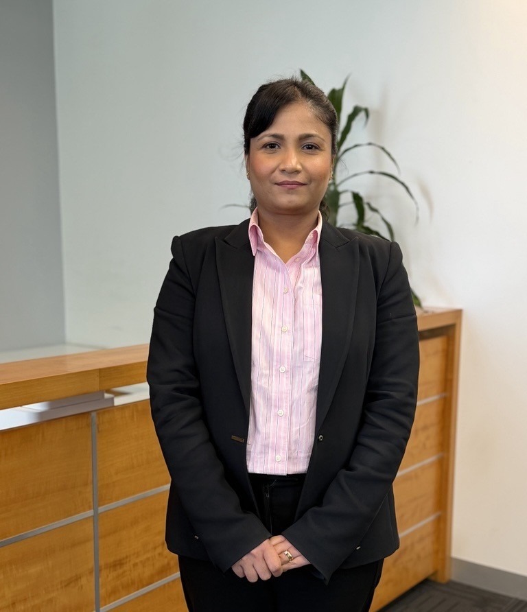 A woman in a black blazer and pink striped shirt stands in an office.