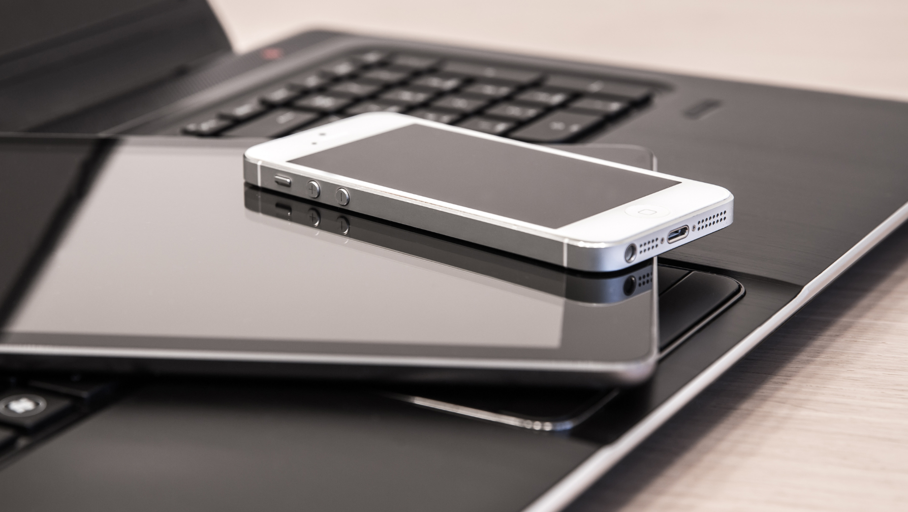 A close-up of a stack of electronic devices, including a smartphone, a tablet, and a laptop. The smartphone is silver with a black screen, the tablet has a black cover, and the laptop has a black keyboard. All devices are placed on a light-colored surface, ready for any SMSF accountant to streamline their workflow.