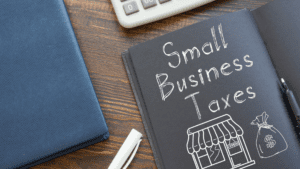 A wooden desk displays a calculator, an open notebook with "Small Business Taxes" written on the right page, a white chalk piece, and a black pen. The left page features a sketch of a store with an "OPEN" sign and a money bag. A blue folder from a Melbourne Accounting Firm is also on the desk.