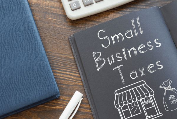 A wooden desk displays a calculator, an open notebook with "Small Business Taxes" written on the right page, a white chalk piece, and a black pen. The left page features a sketch of a store with an "OPEN" sign and a money bag. A blue folder from a Melbourne Accounting Firm is also on the desk.