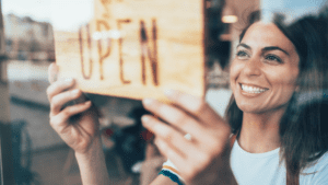 A smiling woman is placing an "OPEN" sign on the glass door of a storefront, her cheerful demeanor radiating through her casual white shirt. The blurred street outside subtly reflects the vibrant energy of a Melbourne accounting firm ready to assist with all your SMSF accountant needs.