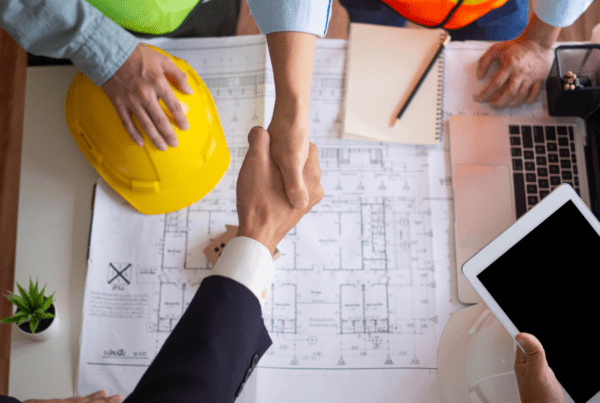 A businessperson and a construction worker shaking hands over a table with construction blueprints, a yellow hard hat, a tablet, and office supplies. Another person, possibly the tax accountant, is also visible, partially in frame, pointing at the blueprints.