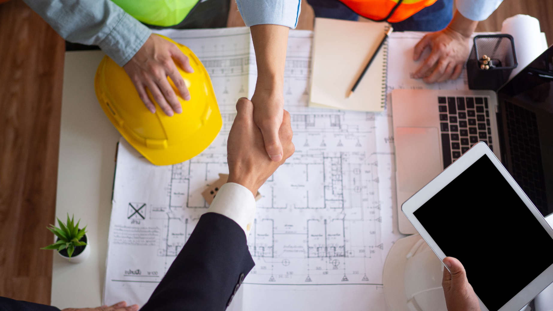 A businessperson and a construction worker shaking hands over a table with construction blueprints, a yellow hard hat, a tablet, and office supplies. Another person, possibly the tax accountant, is also visible, partially in frame, pointing at the blueprints.