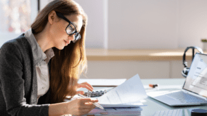 A woman with long hair and glasses works at a desk. She is looking at documents and using a calculator. A laptop displaying an invoice from a Melbourne accounting firm is open in front of her, along with pens and a stack of papers. The bright, well-lit workspace suggests she might be a tax accountant.